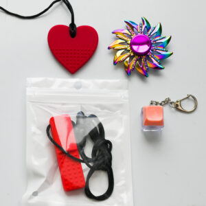 magenta heart chew necklace, rainbow fidget spinner, red bar chew necklace, and a coral colored keyboard fidget keychain
