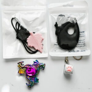 a leaf chew necklace, teardrop chew necklace, rainbow fidget spinner, and paw keyboard fidget keychain together on a white reflective surface