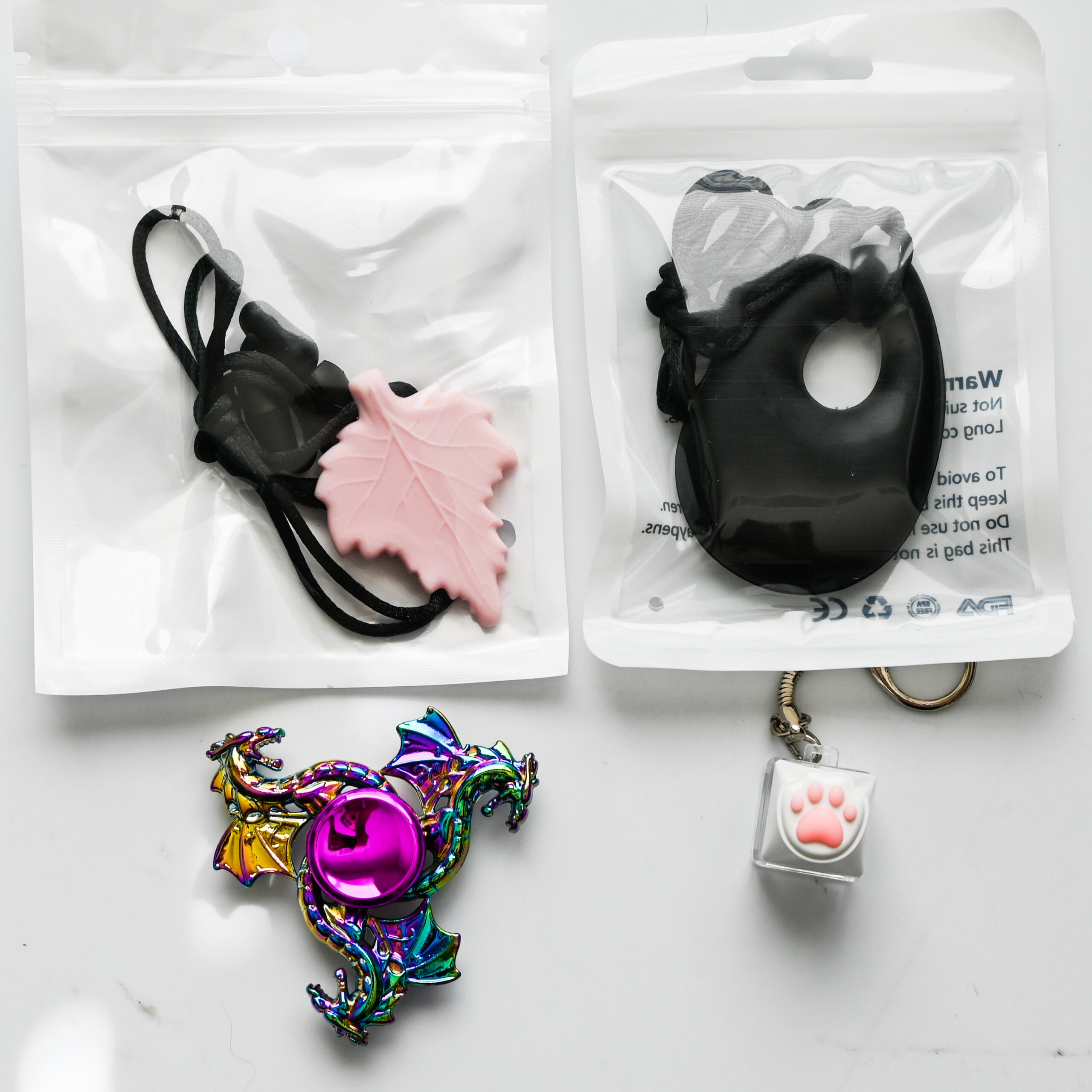a leaf chew necklace, teardrop chew necklace, rainbow fidget spinner, and paw keyboard fidget keychain together on a white reflective surface