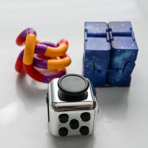purple yellow red twisty fidget toy, blue galaxy colored galaxy infinity fidget cube, and a shiny silver push button fidget cube on a white reflective surface