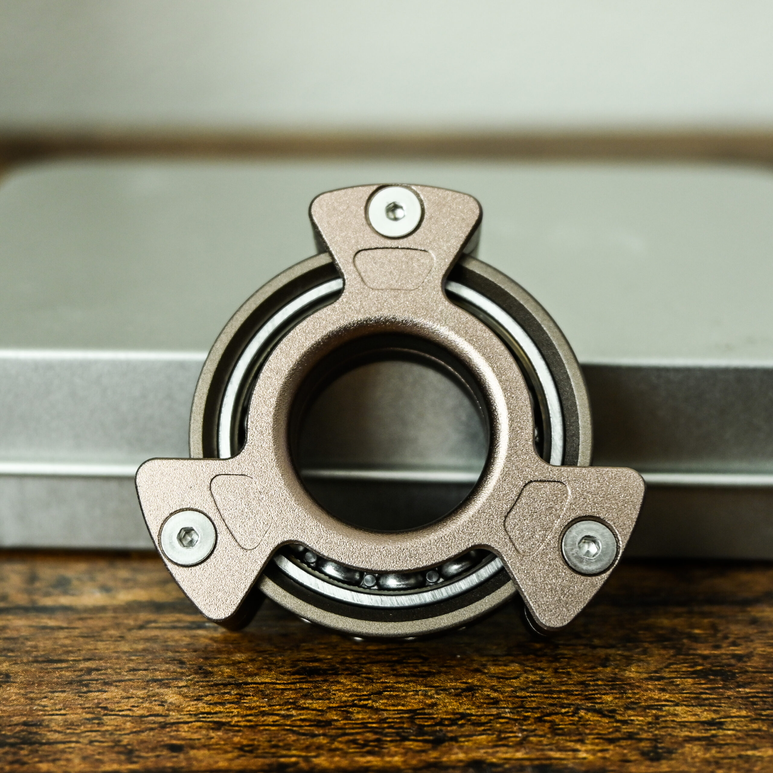 grey fingertip fidget spinner with 3 notches propped up against a thin silver metal box on a wood table.