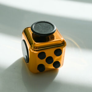shiny copper metal color fidget with buttons, turn wheel, joystick, switches. It's sitting in a block of light with alternating blocks of shadows and light behind it and a copper rainbow reflection on the shadows