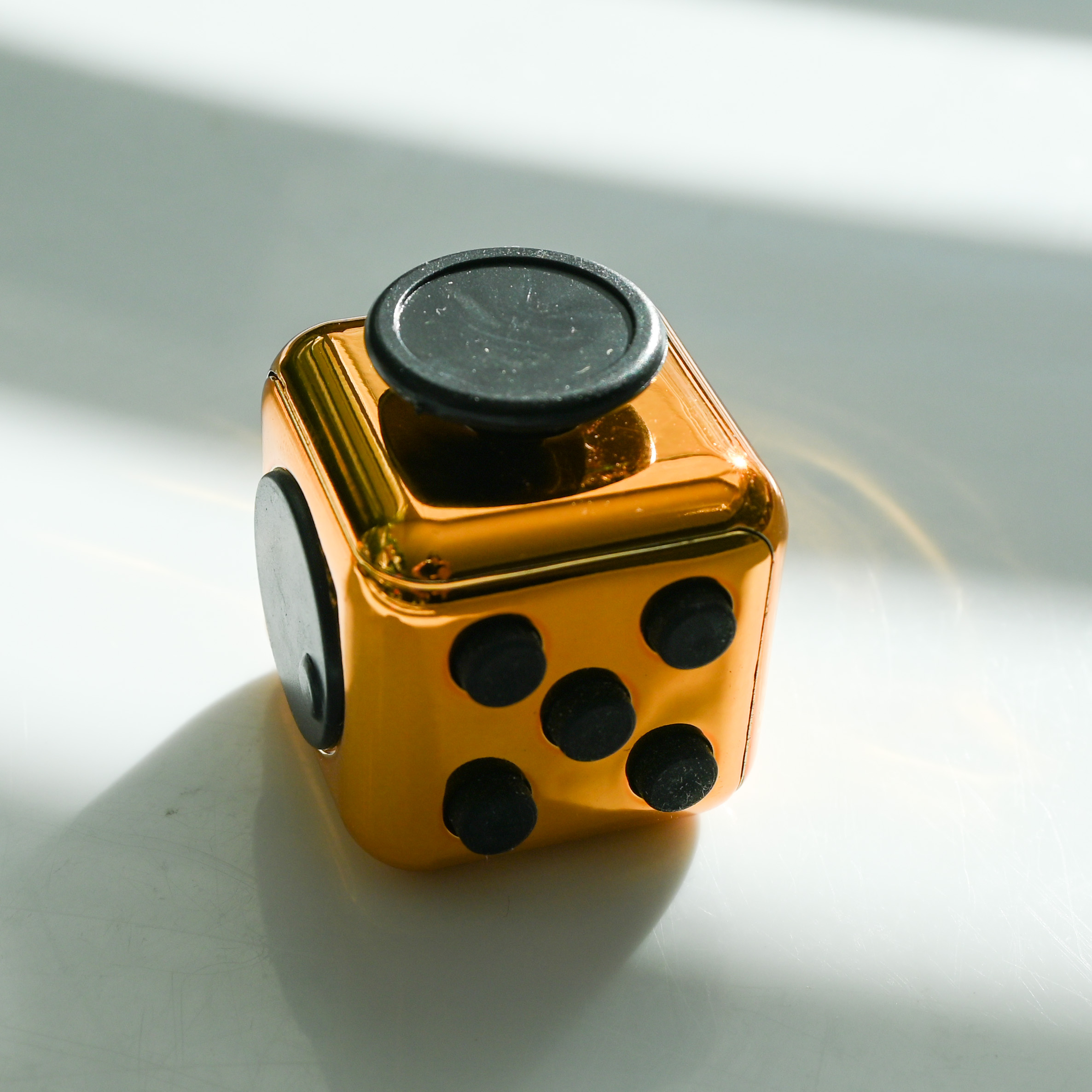 shiny copper metal color fidget with buttons, turn wheel, joystick, switches. It's sitting in a block of light with alternating blocks of shadows and light behind it and a copper rainbow reflection on the shadows