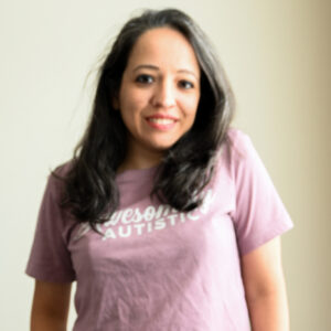 I'm smiling and standing in front of a blank wall where the sunlight through the window made a slight yellow haze on the background. I'm wearing an "Awesomely Autistic" pink t-shirt and blue jeans with my hands in my pockets.
