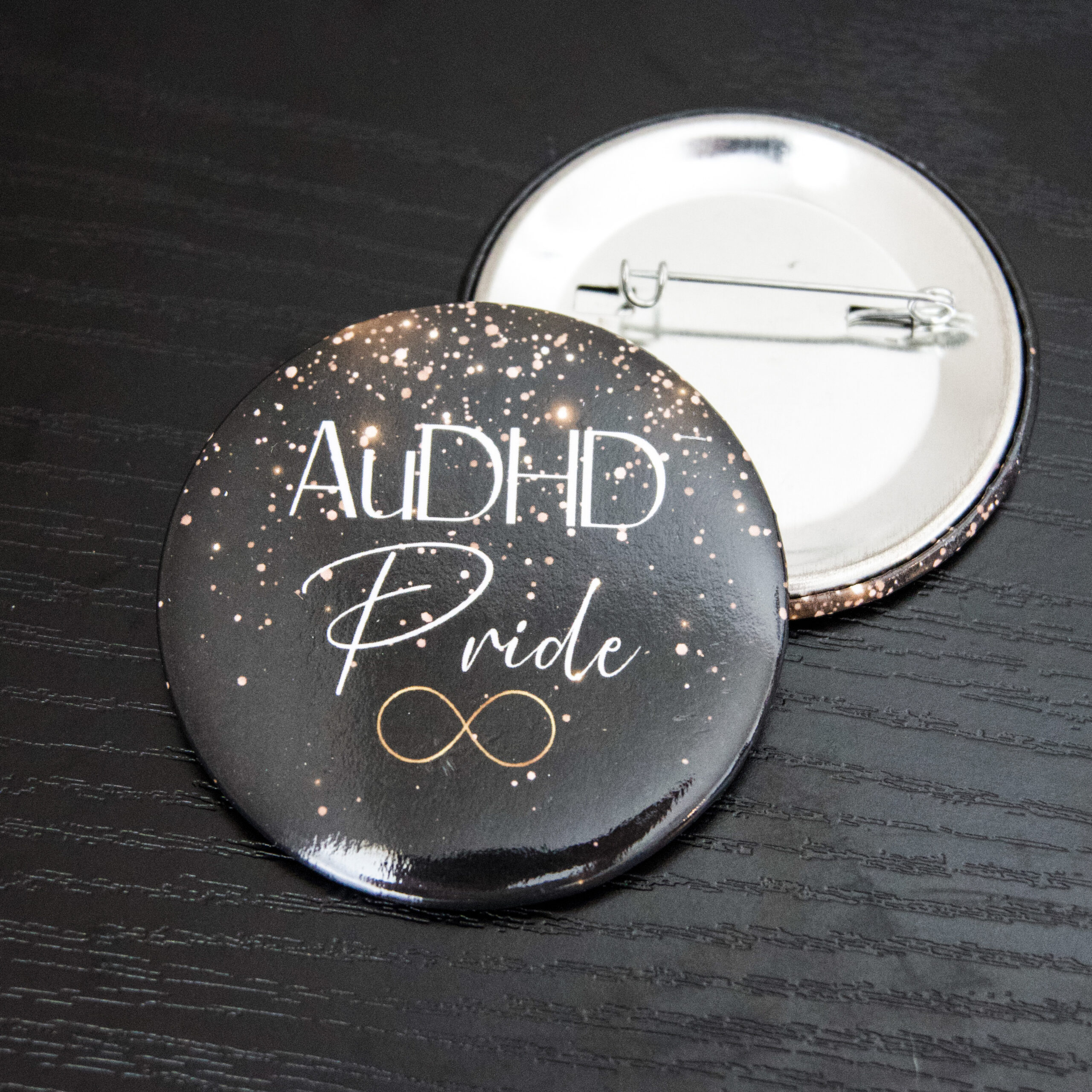 black pin with gold glitter design and the text "AuDHD Pride" and a gold infinity symbol underneath.