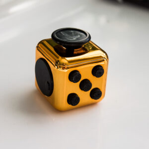 shiny copper metal color fidget with buttons, turn wheel, joystick, switches. It's sitting in a block of light with alternating blocks of shadows and light behind it and a copper rainbow reflection on the shadows