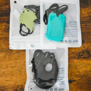 Green leaf pendant chew necklace, turquoise dog tag chew necklace, and black teardrop chew necklace inside the bag packaging on a wood table